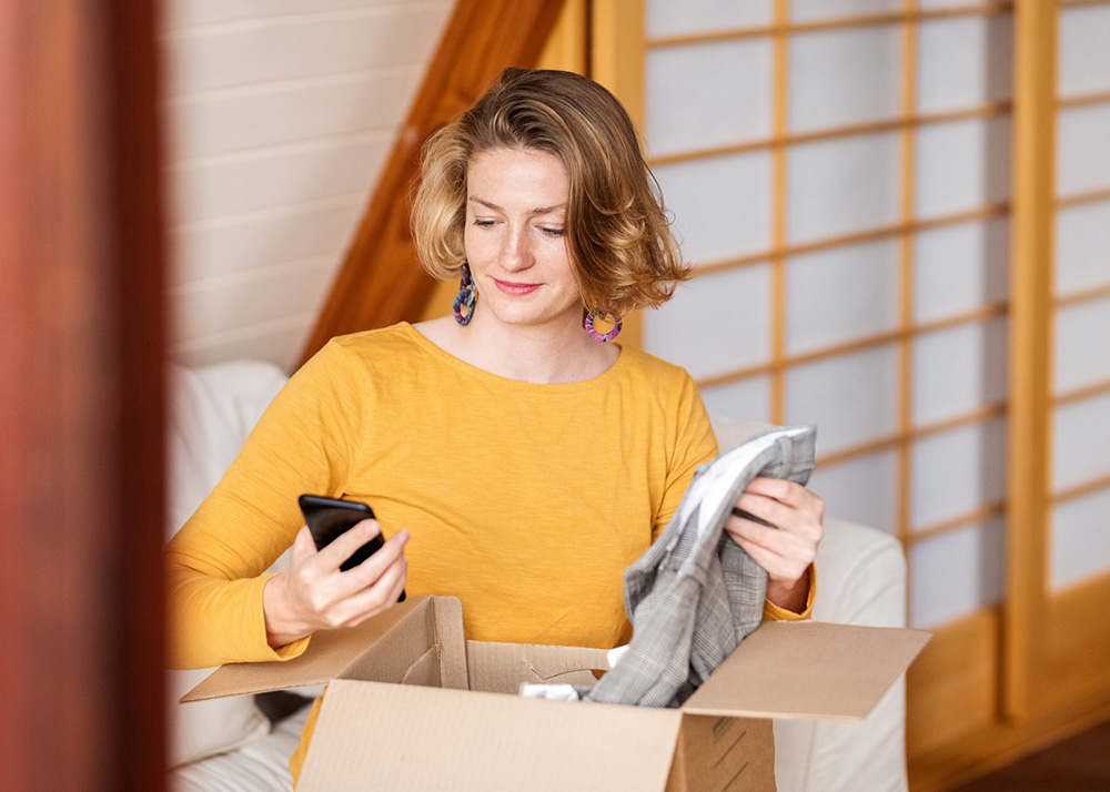 Young woman looking at her phone while unboxing a package from online retail delivery to check if the products are correct