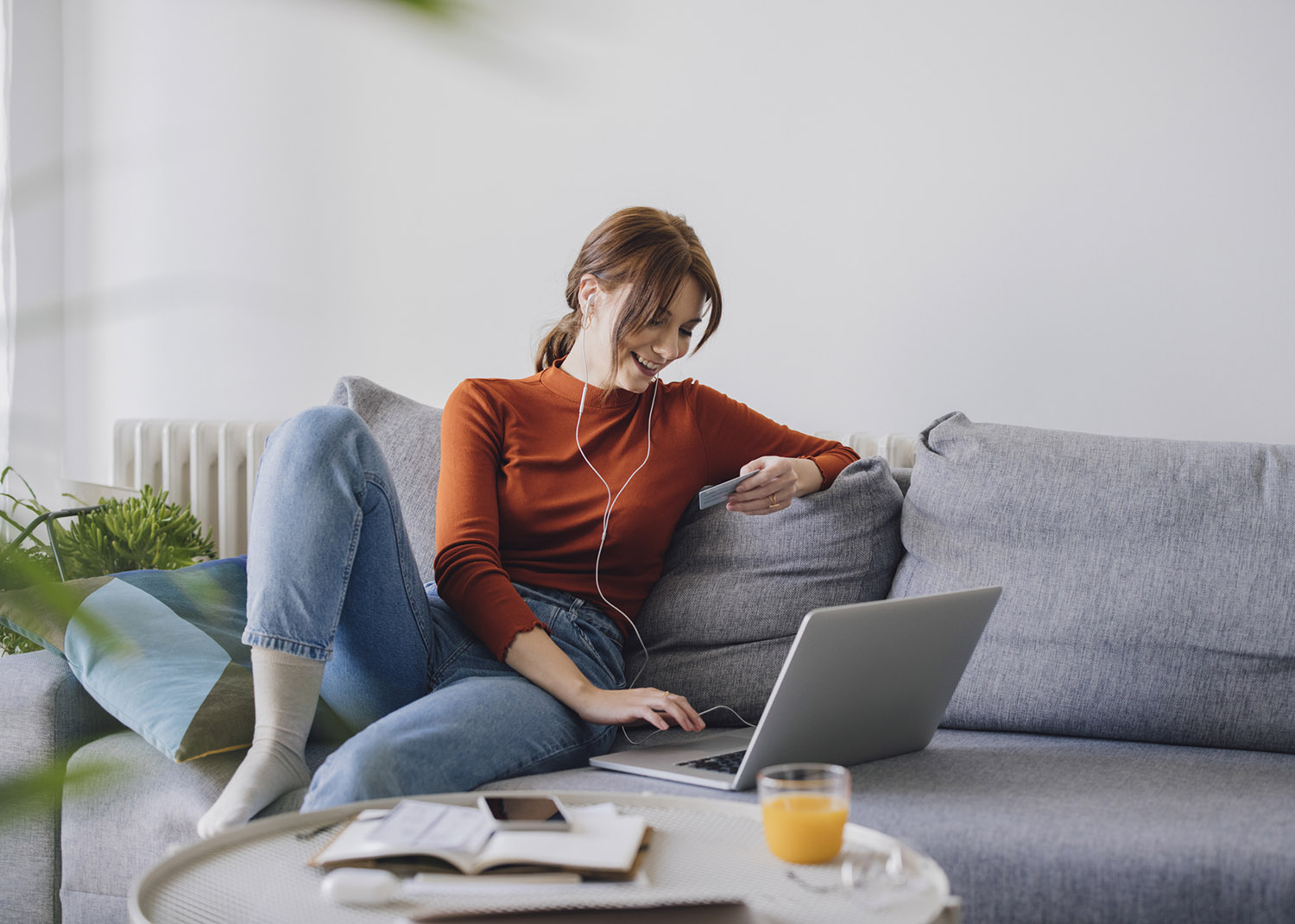 A beautiful young Caucasian businesswoman wearing a red long sleeve shirt and jeans holding a credit card and using a laptop for online shopping while sitting comfortably on a sofa at home.