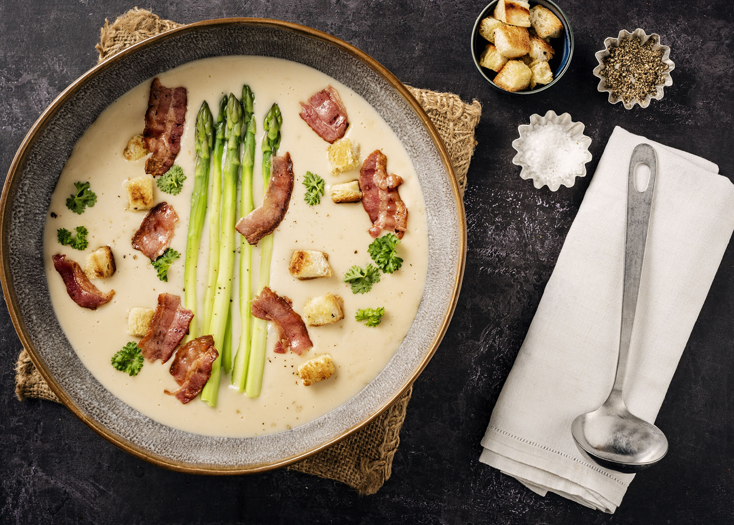 Overhead view of a serving bowl full of loaded creamy potato soup with fresh asparagus stems, croutons and pieces of crispy bacon. Colour, horizontal with some copy space.