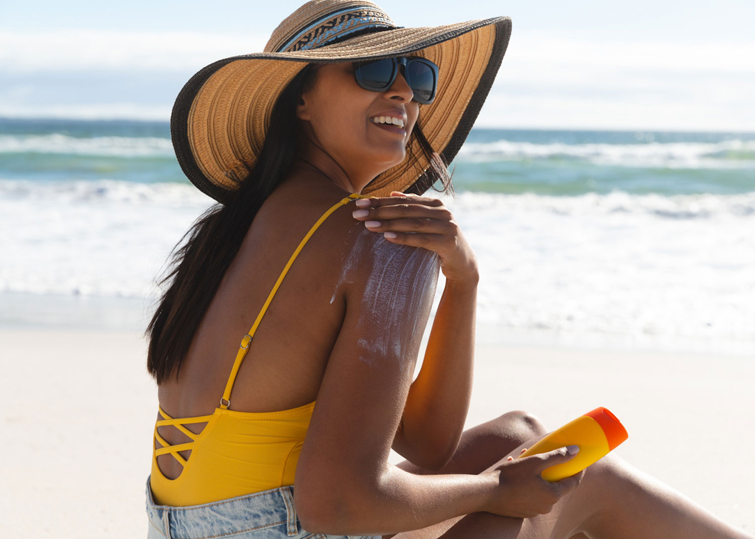 femme avec costume de bain jaune et chapeau se met de la crème solaire 