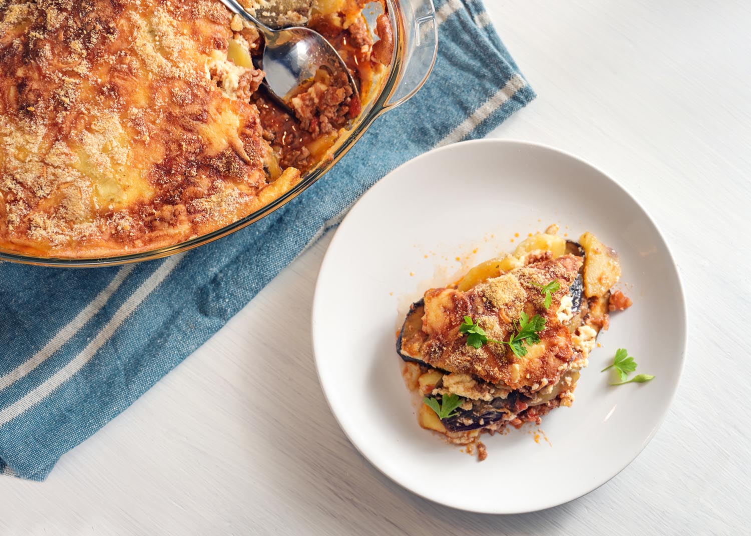 Moussaka baked in a glass casserole and served on a white plate, traditional Greek dish of eggplants, potatoes, minced meat and tomatoes, copy space, high angle view from above, selected focus