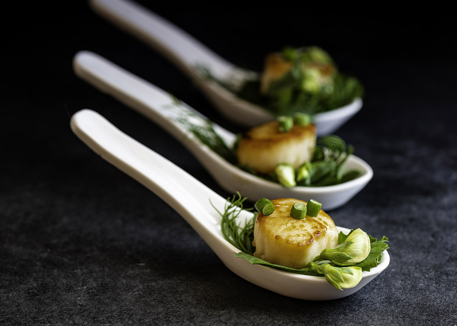 Scallops seared in a cast-iron pan with butter and garlic scapes. Served in ceramic spoons as an appetizer on a bed of pea tendrils and blossoms.
