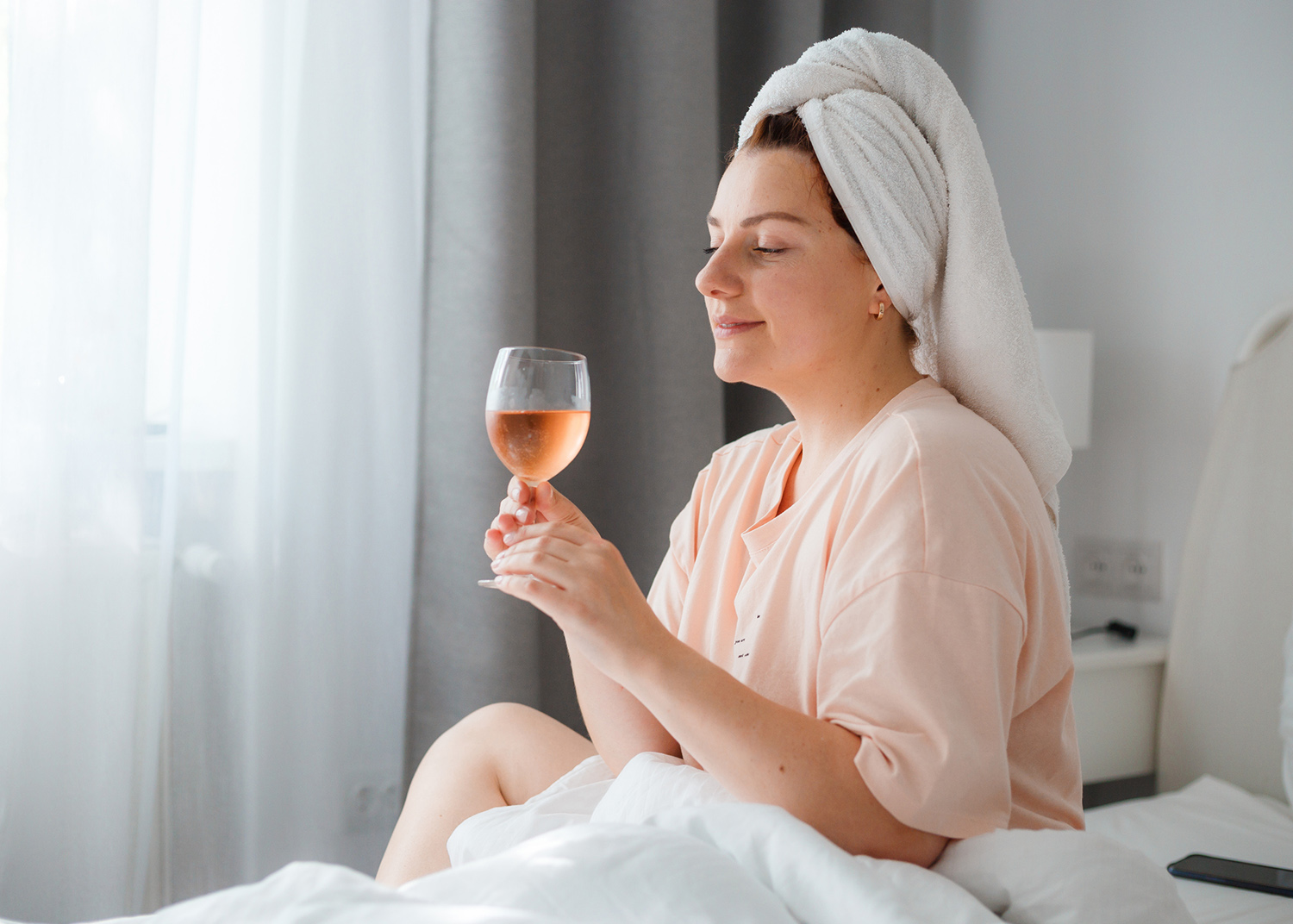 Beautiful young woman after shower with glass of wine in bedroom. Staying at home in isolation during quarantine lockdown.
