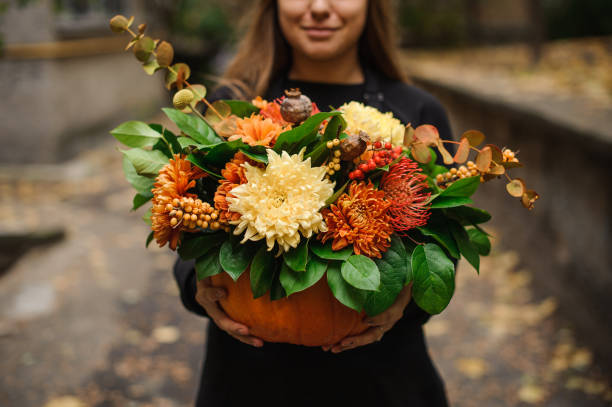 Smiling woman in black dress holding a beautiful pumpkin with beautiful autumn flowers on the outdoors background