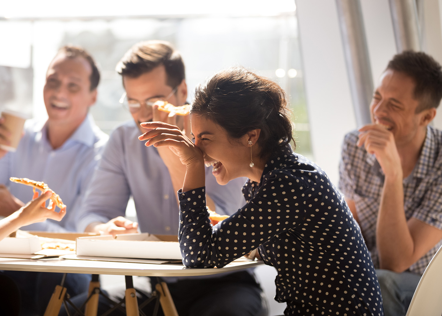 Indian woman laughing at funny joke eating pizza with diverse coworkers in office, friendly work team enjoying positive emotions and lunch together, happy colleagues staff group having fun at break