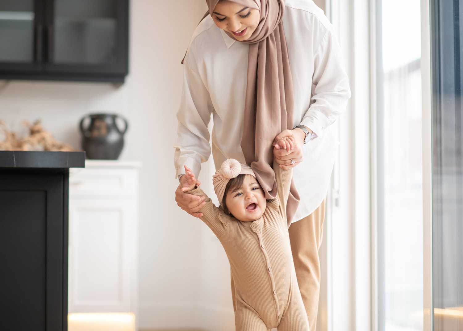 A Muslim Mother holds her daughters arms as she teaches her how to walk.  They two are dressed in muted neutral tones and traditional headdress as they walk around their living room.  The baby girl has a huge smile on her face as she walks with her Mother.