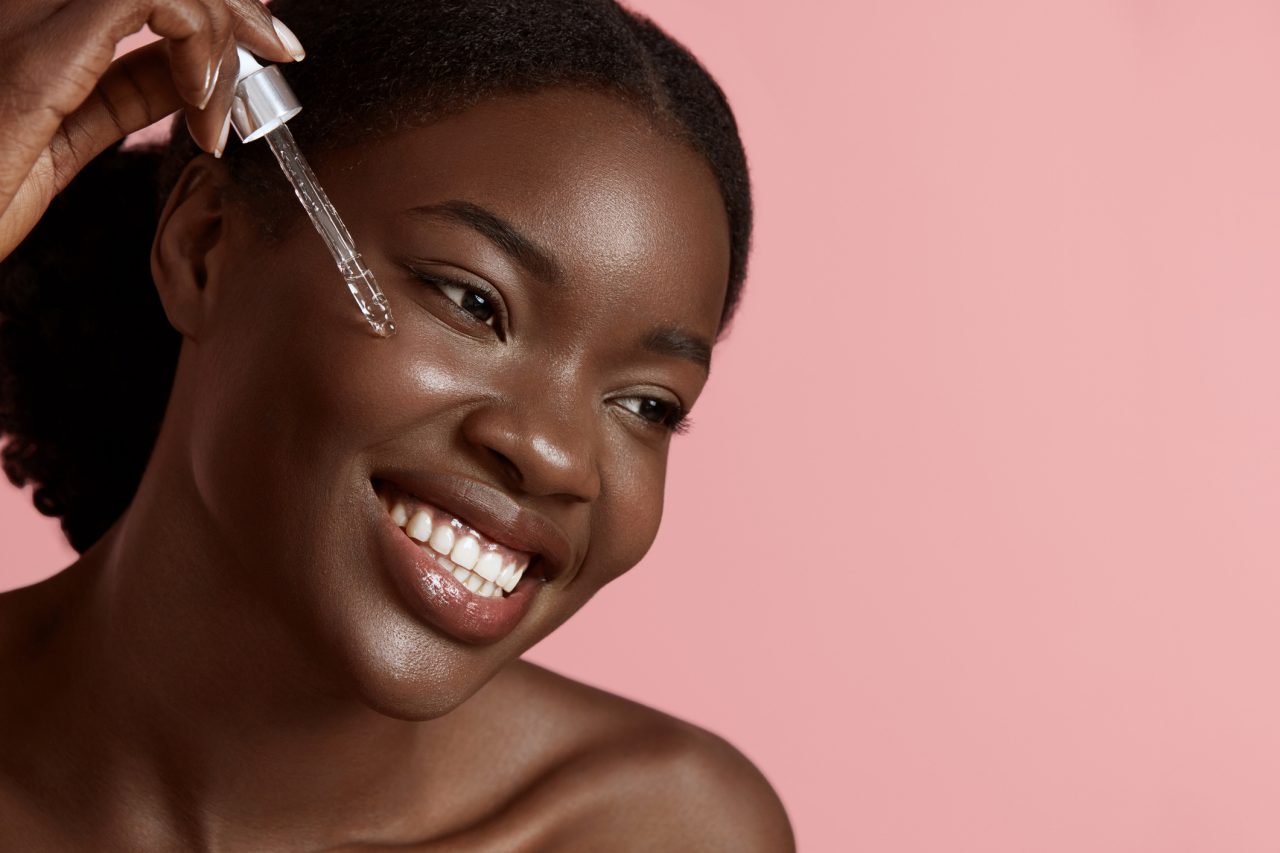 Portrait close up of beautiful black girl dropping serum collagen moisturizer on face. Smiling young woman. Concept of face skin care. Isolated on pink background. Studio shoot