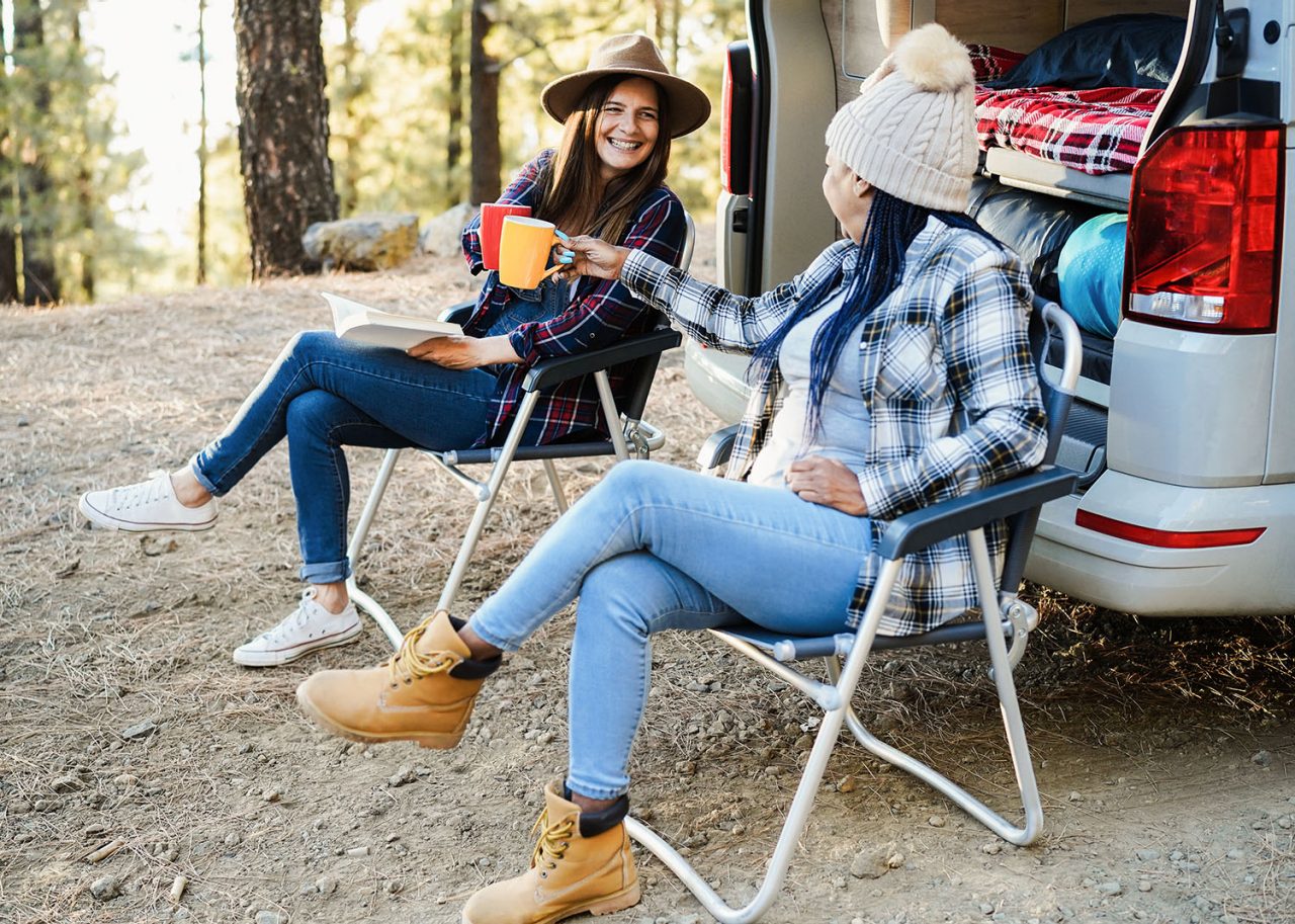 Multiracial women friends having fun camping with camper van cheering with coffee outdoor - Focus on hand holding cup