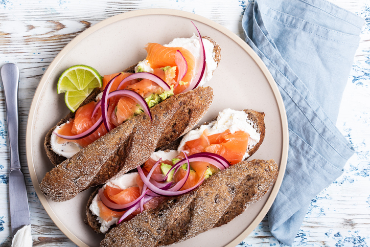 Whole wheat baguette sandwiches with cured salmon, curd cheese, red onion, avocado and lime on rustic table viewed from above