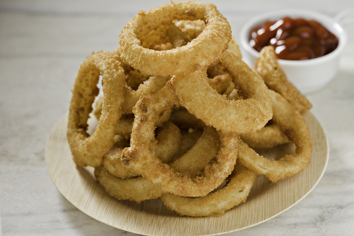 An extreme close up of a small bamboo plate with a stack of golden brown onion rings and a small container of ketchup in  the foreground. Shot on a white marble surface.