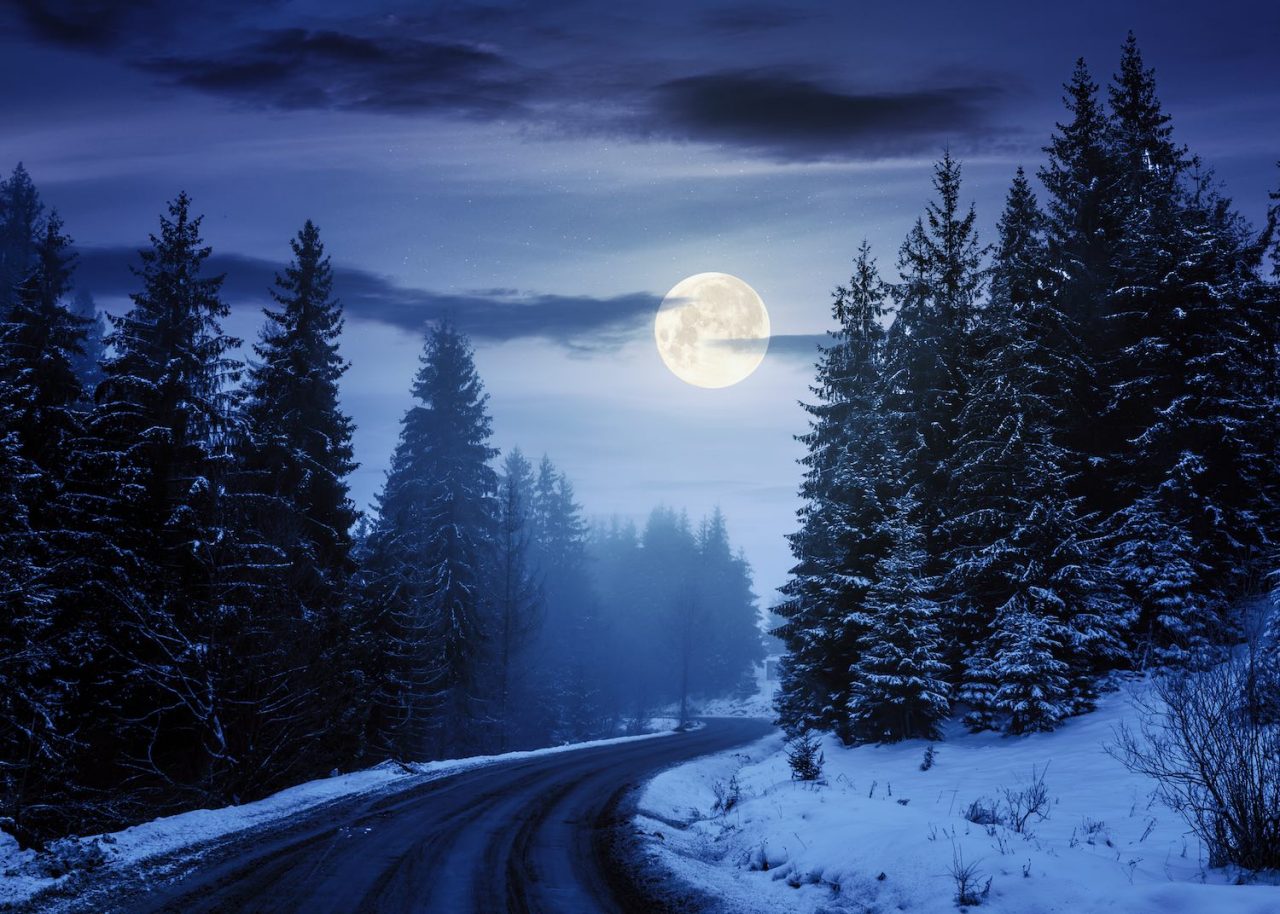 country road through forest at night. misty winter weather in full moon light. snow on the roadside. cloudy sky