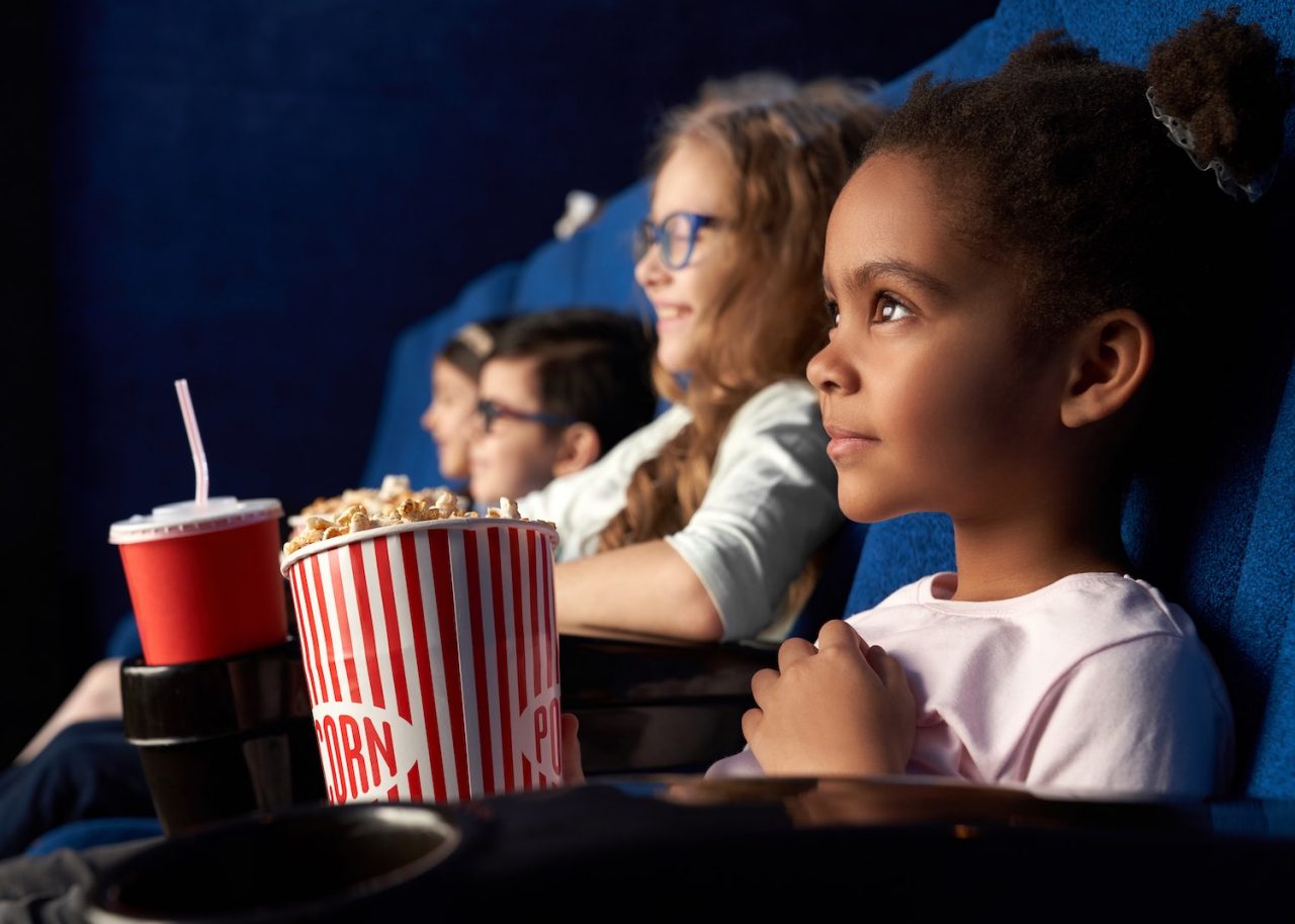 Beautiful african girl with funny hairstyle watching excited movie in cinema. Wonderful little child sitting with friends, eating popcorn and smiling. Concept of entertainment and enjoyment.
