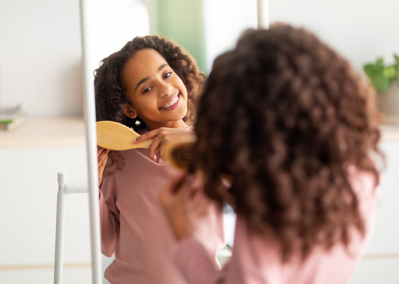 Morning beauty routine. Happy african american girl combing her curly hair with wooden comb, getting ready for school or walk, standing near mirror and smiling, over shoulder view