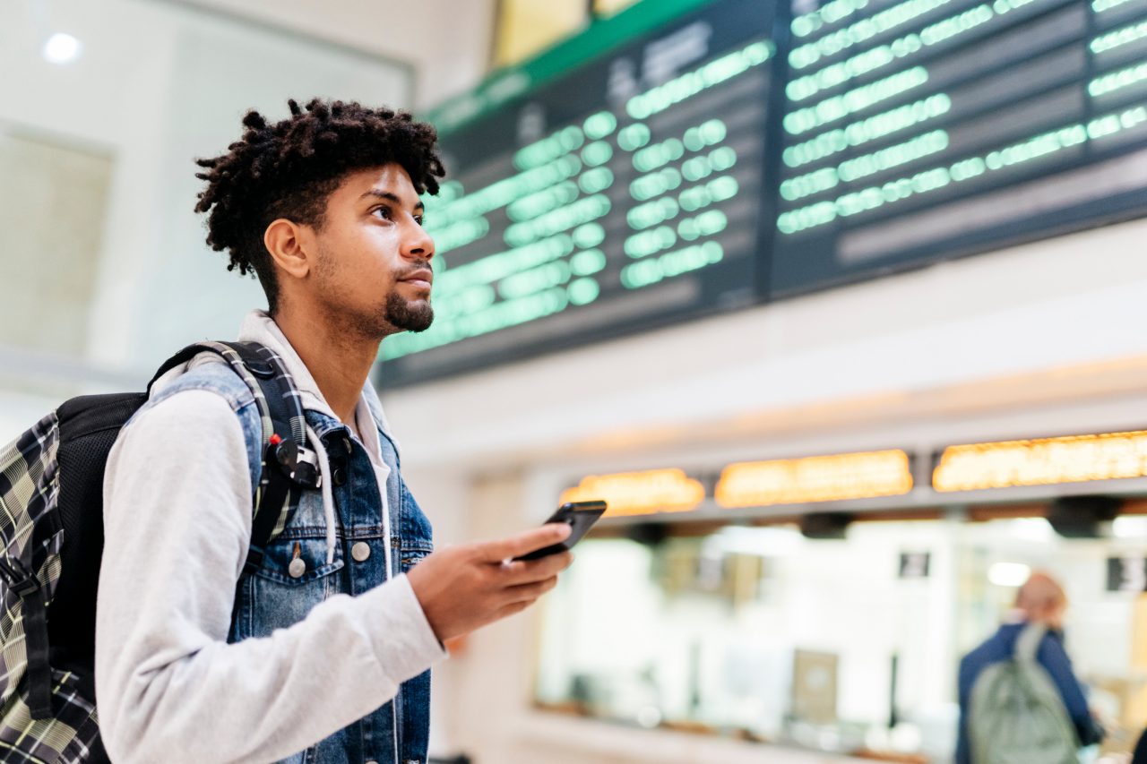 young afro man holding a mobile phone looking at the departures of planes in an airport