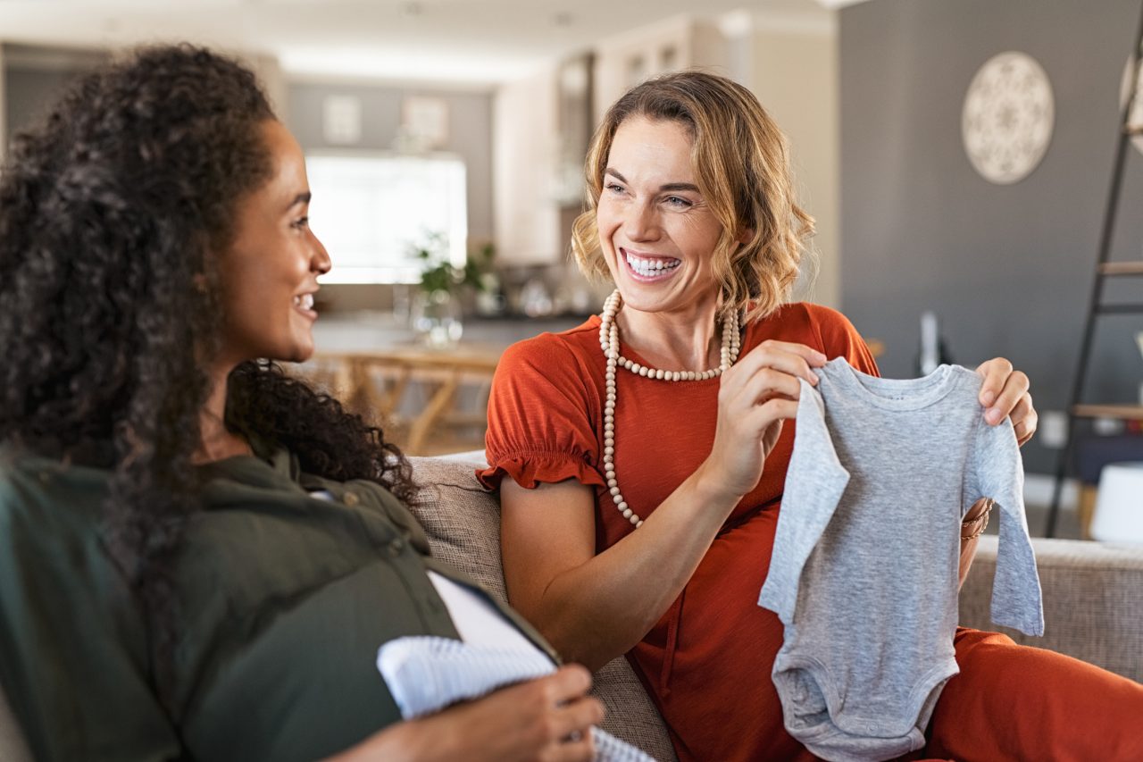 Happy pregnant woman looking at baby clothes with joyful african friend. Laughing pregnant friends sitting on sofa and looking at baby shower gifts. Excited two expecting girls checking baby clothes for new baby.