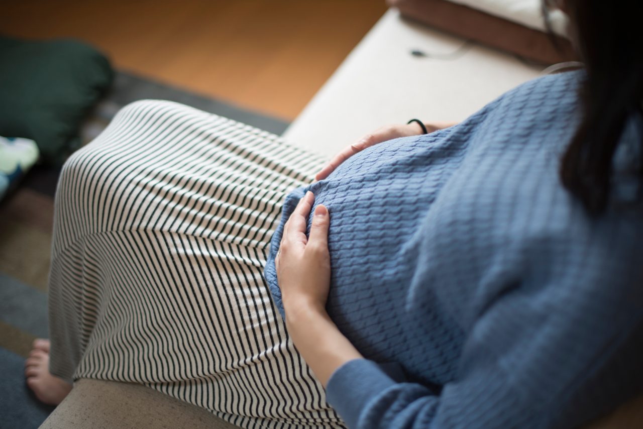 A woman in 9th months of pregnancy pats her belly gently at living room.