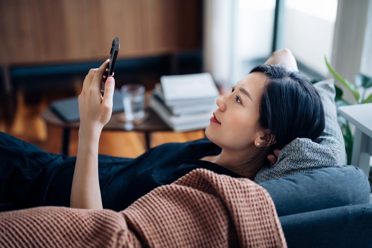 Carefree young Asian woman using smartphone while relaxing on sofa at cozy home. Technology in everyday life. Lifestyle and technology