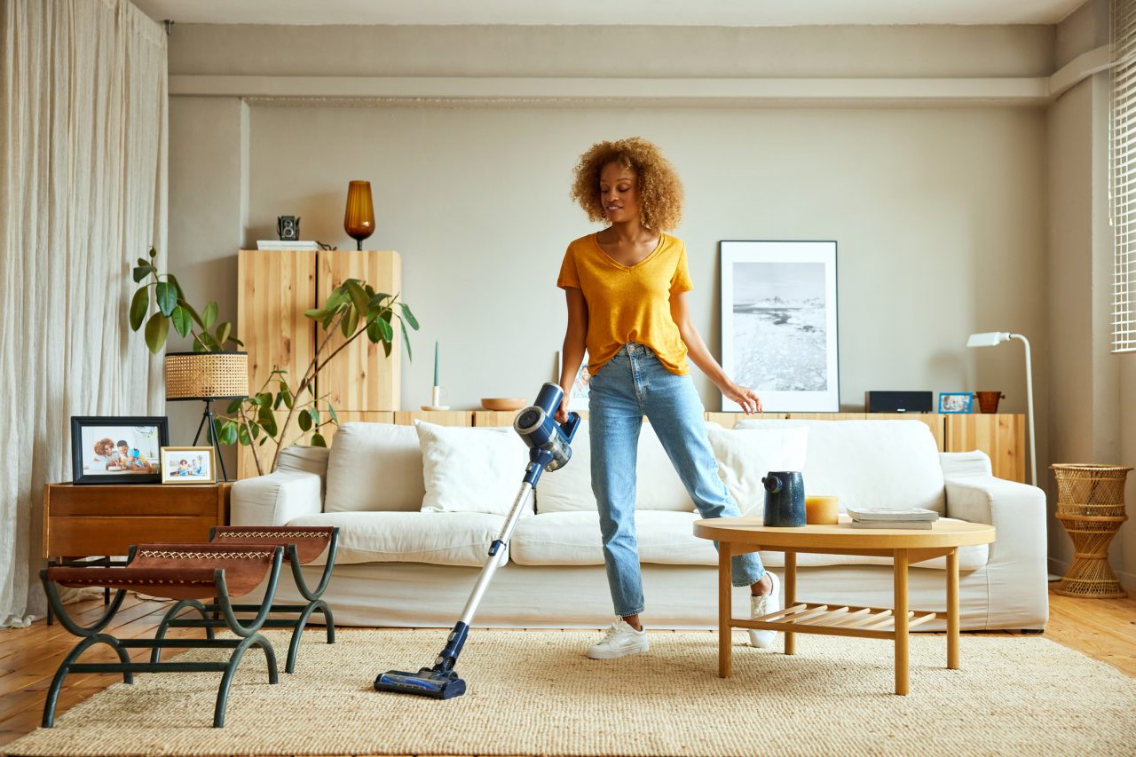 Smiling young woman cleaning carpet with vacuum cleaner at home. Female is doing housework in living room. She is standing against sofa.