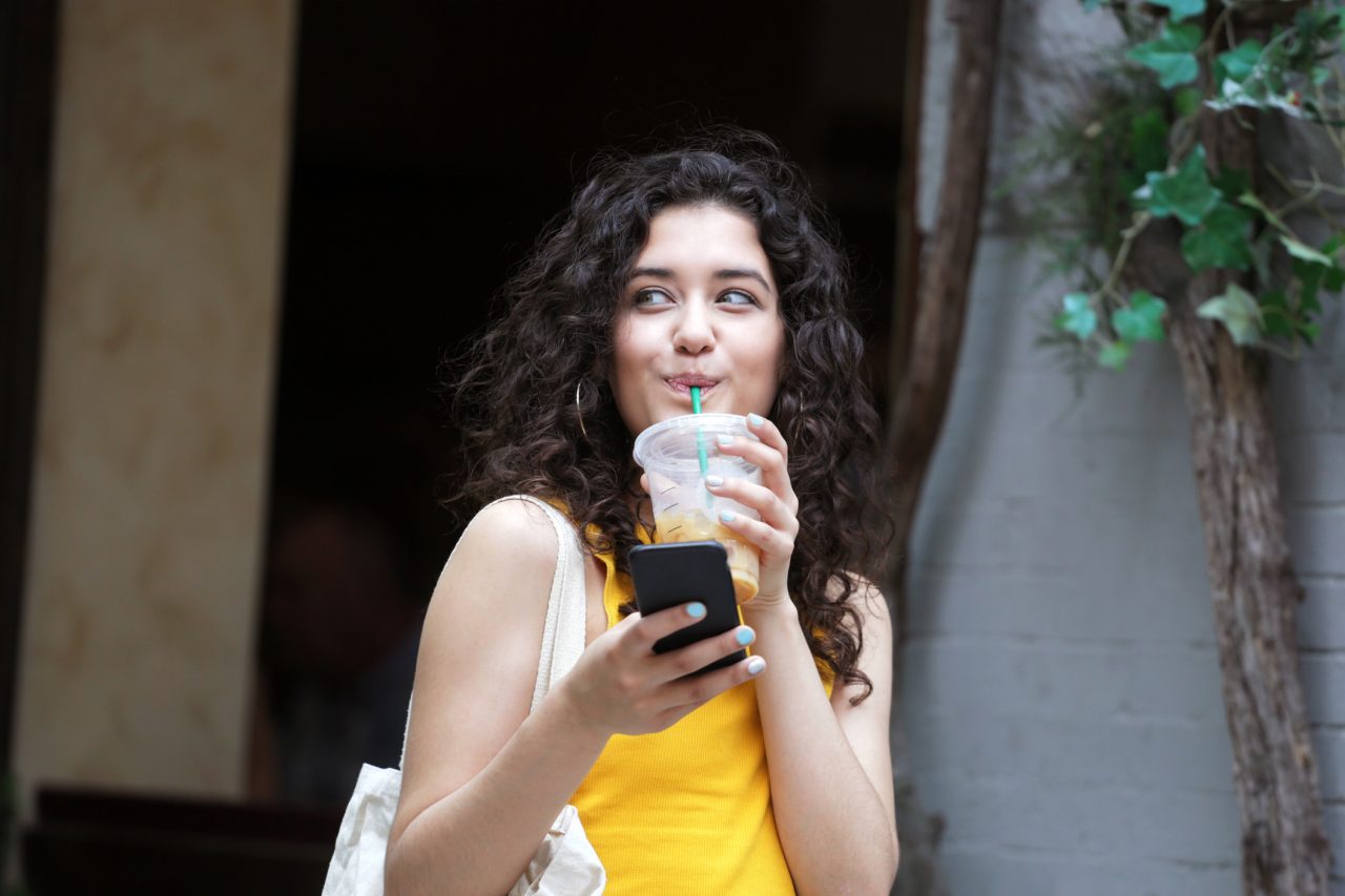 A LatinX female millennial smiles while holding a cell phone and drinking an iced coffee, on a summer day in New York City.