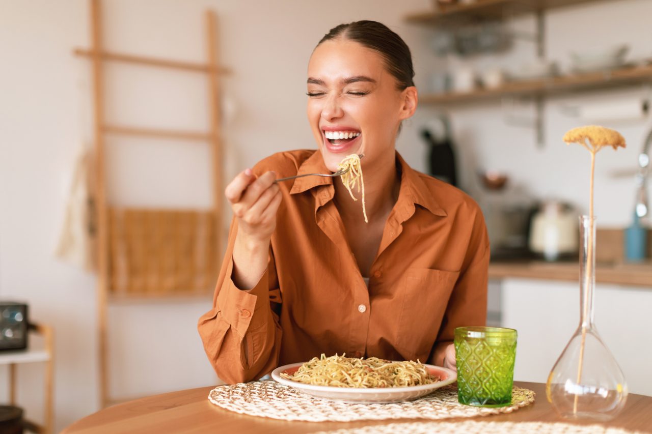 Italian inspiration. Happy young european woman enjoying freshly cooked pasta, holding fork and smiling, sitting in cozy kitchen interior, free space