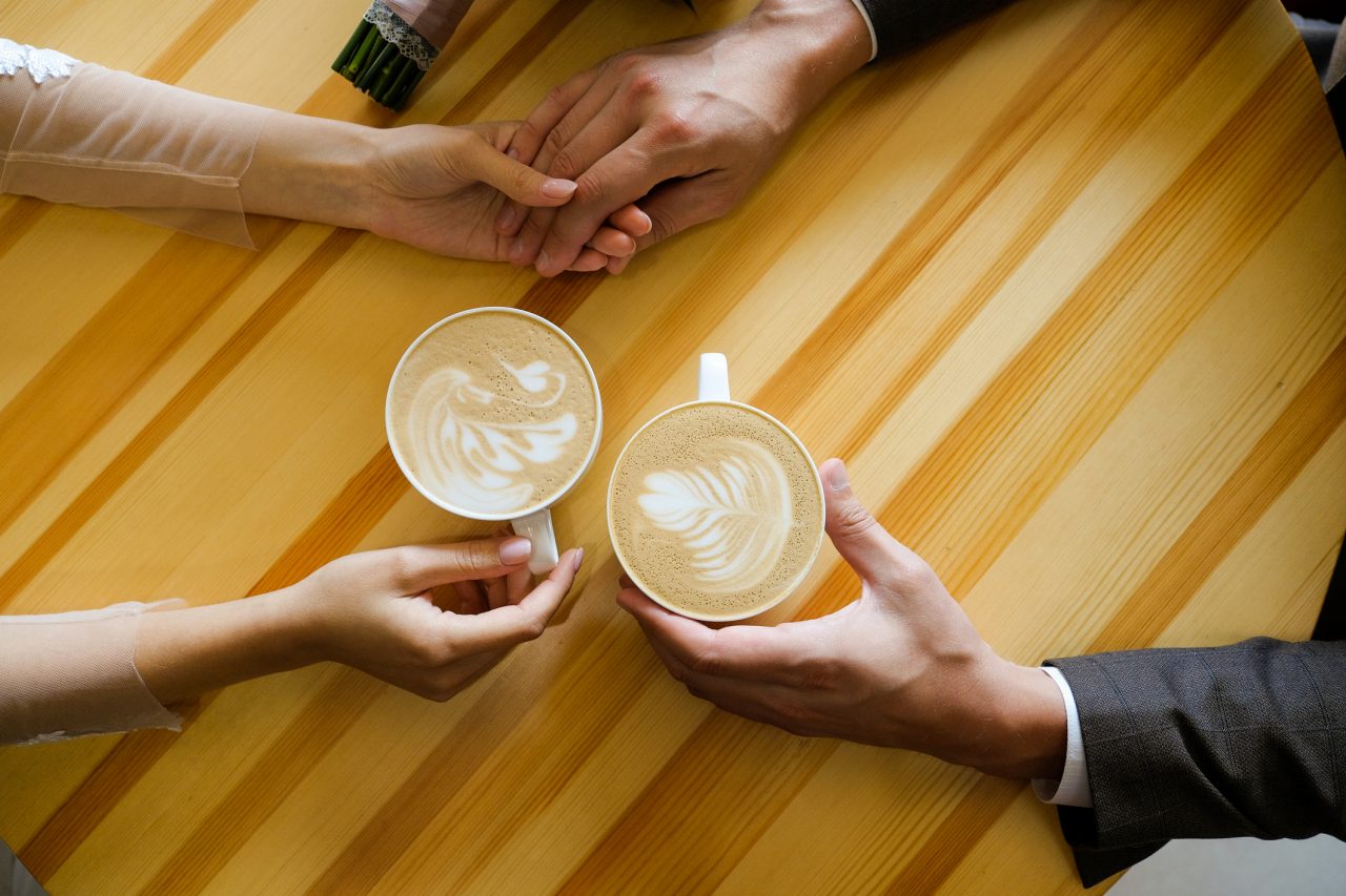 A man and a woman holding hands in a cafe, drinking coffee. Lovers' hands on the background of a wooden table. Breakfast or lunch at the restaurant. Engagement, a guy holding his girlfriend's hand tightly. Romantic meeting, Valentine's Day.