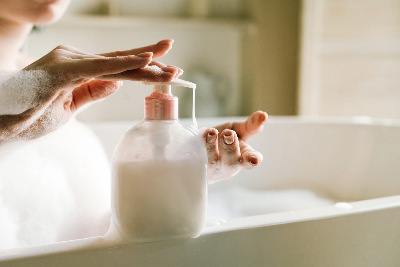 Dispenser with white liquid soap or shower gel, for intimate hygiene. Woman's hands pushing a button. White toilet soap in female hands against the background of a fragrant foam bath. Woman in white terry bathrobe washing her hands with soap. Relaxation and beauty treatments. Natural beauty, daily skincare routine. Moisturizing, cleansing