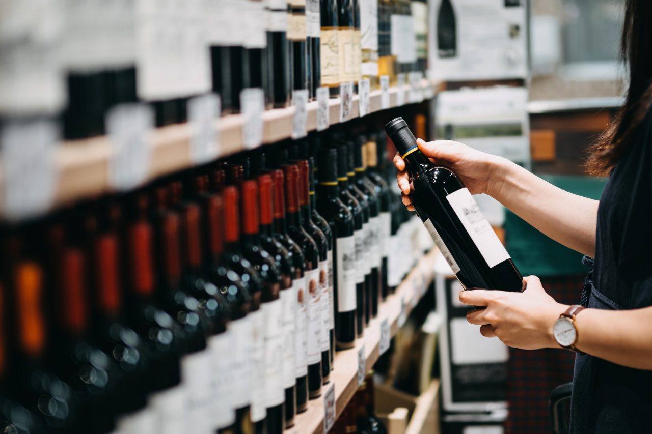 Close up of young Asian woman walking through supermarket aisle and choosing a bottle of red wine from the shelf in a supermarket