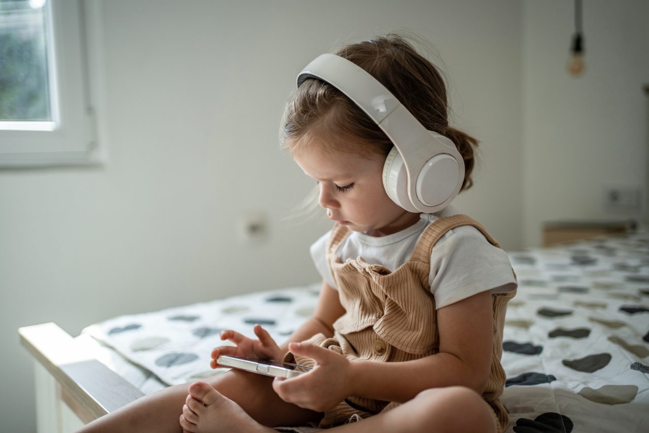 Happy little girl enjoys watching cartoon on mobile phone at home, she has wireless headphones on her head.