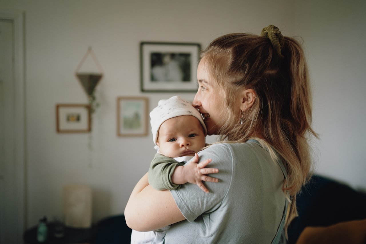 Mum with long blonde hair holding baby who is looking at camera with fingers outstretched