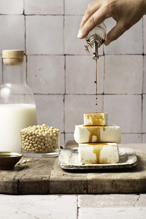 large group of plant based products on wooden board with copy space, soy milk in glass bottle, soy beans, tofu cubes, soy sauce and miso sauce, human hand pouring soy sauce on tofu cubes, copy space.