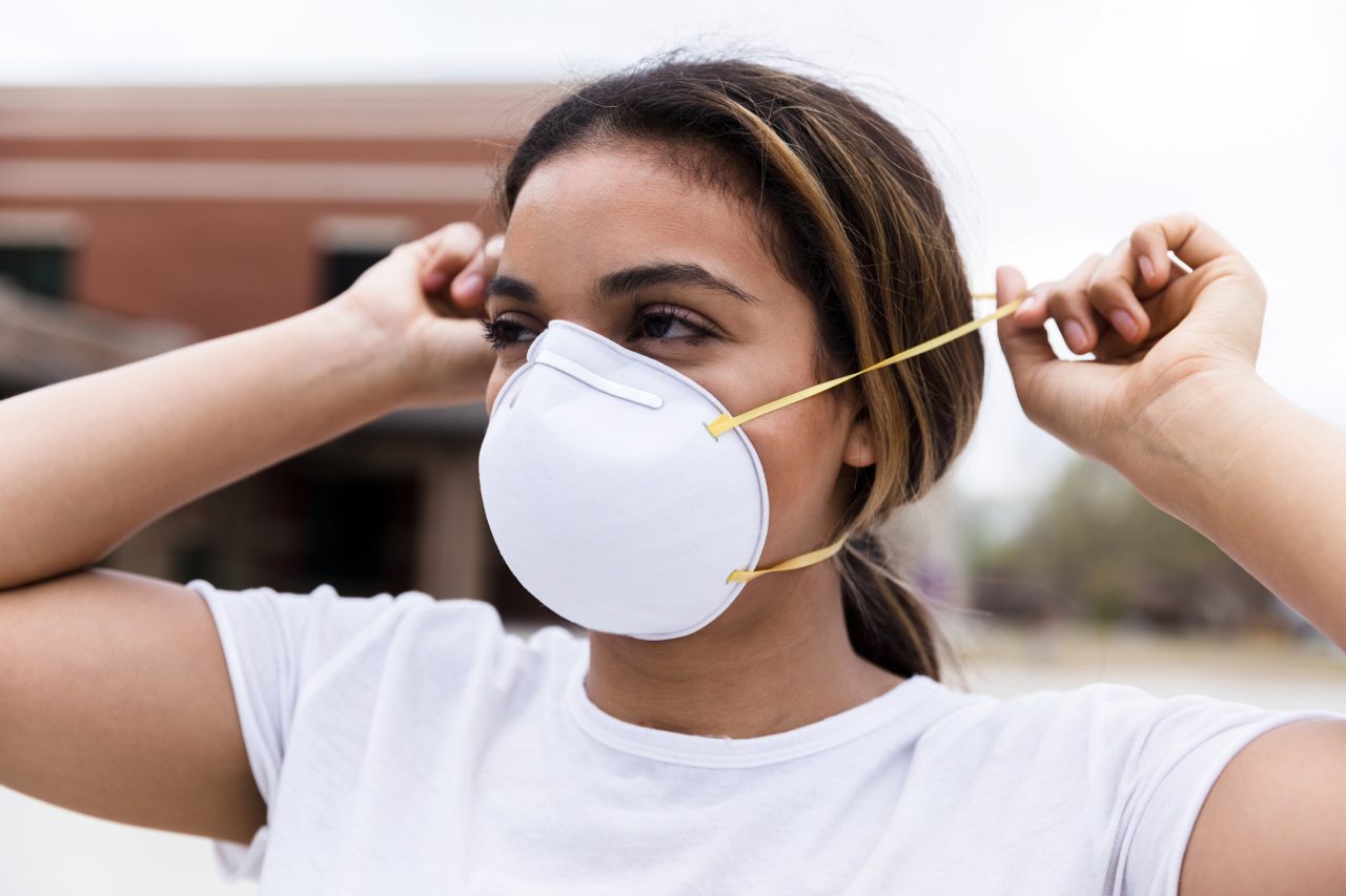 During a pandemic, a mid adult woman protects herself by placing an N95 face mask over her nose and mouth. She is standing outdoors.