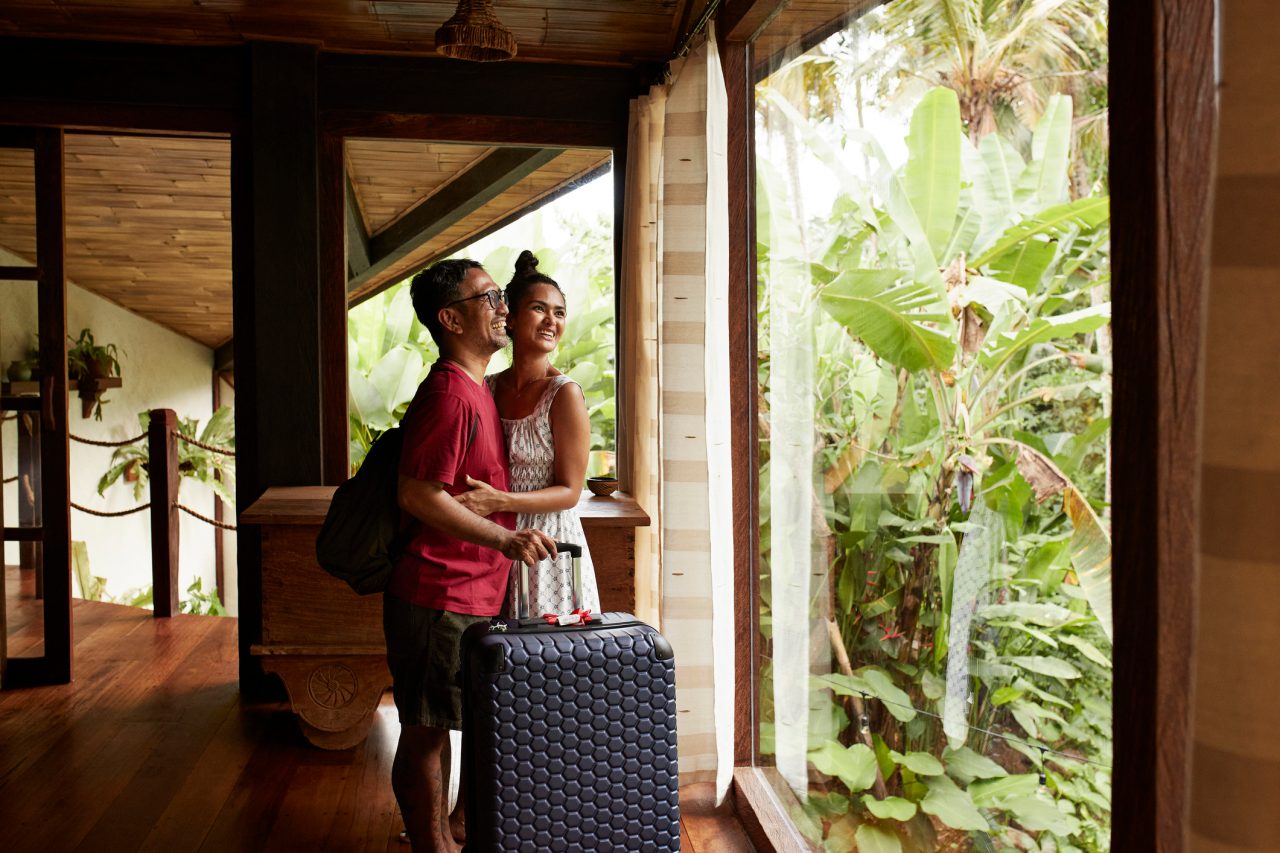 Happy man with luggage standing by woman near window during vacation