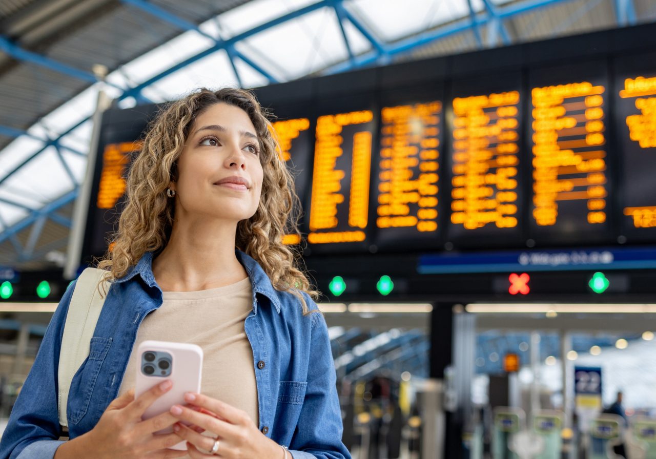Young woman at the train station checking the departure board and using her phone - travel concepts